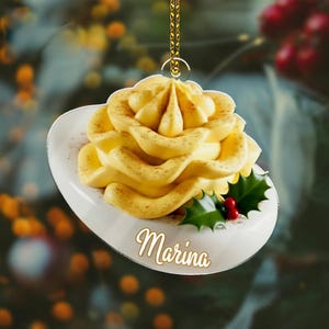 May include: A decorative Christmas ornament shaped like a dessert, featuring a swirl of yellow frosting dusted with brown powder, on a white plate. The ornament includes a sprig of holly with red berries and the name "Marina".