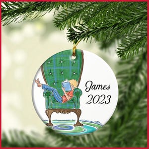 May include: Round Christmas ornament featuring a cartoon illustration of a person reading in a green armchair. The ornament has the name "James" and the year "2023" printed on it. The ornament is hanging from a gold string.