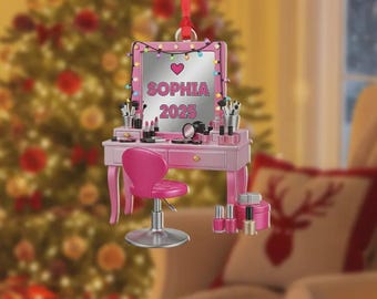 May include: A pink vanity Christmas ornament with a mirror, lights, and a heart. The mirror reads "SOPHIA 2025." The vanity has a matching chair, makeup, and nail polish. A red ribbon is attached for hanging.