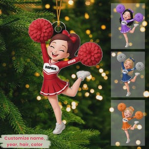 May include: A red and white cheerleader ornament with the name "Sophia" and the year "2025" on the uniform. The ornament is holding red pom-poms and is hanging on a Christmas tree. Additional cheerleader ornaments in purple, blue, and orange are also shown.