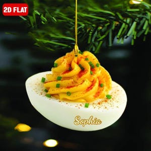 May include: A deviled egg Christmas ornament with a yellow filling, sprinkled with paprika and green chives. The white eggshell has the name "Sophia" on it. The ornament is hanging from a gold string, with the text "2D FLAT" in the upper left corner.