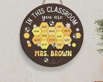 May include: A round wooden classroom sign with the text "IN THIS CLASSROOM you are" and a honeycomb design with positive words. The sign includes the name "MRS. BROWN" and bee and flower accents.
