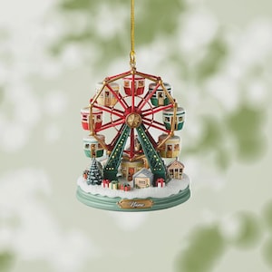 May include: A festive Christmas ornament featuring a miniature Ferris wheel with red, green, and gold accents. The ornament includes small houses, a Christmas tree, and gift boxes, all set on a snowy base. A gold string is attached for hanging.