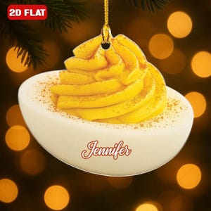 May include: A 2D flat Christmas ornament shaped like a deviled egg. The egg white is white, and the filling is yellow with a dusting of spice. The name "Jennifer" is written in cursive on the egg. The ornament hangs from a gold string.