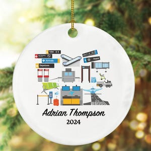 May include: White ceramic ornament with a gold hanging cord. The ornament features an airport-themed design with illustrations of airplanes, baggage, and terminal signs. The name "Adrian Thompson" and the year "2024" are printed in black script.