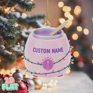May include: A pink Christmas ornament shaped like a wax warmer, with the words "CUSTOM NAME" in purple. It has a purple dial and is strung with colorful lights. A wooden stick rests inside the warmer.