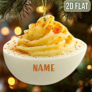 May include: A deviled egg-shaped Christmas ornament with a yellow filling and a dusting of paprika. The ornament has the word "NAME" printed on the front and the text "2D FLAT" on the top right.
