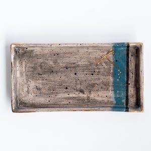 Könnte beinhalten: Ein rechteckiges Keramik-Serviertablett mit einem gesprenkelten grauen Überzug und einem blauen und schwarzen Streifenmuster. Das Tablett hat einen goldenen Akzent in Form einer Libelle.