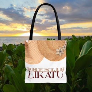 May include: A white tote bag with a black handle, featuring a straw hat design and the text "DRENLIK LIKATU" in brown. The bag is set against a backdrop of a sunset over the ocean, with green foliage in the foreground.