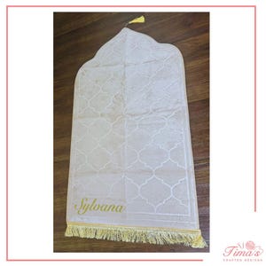 May include: A light beige prayer rug featuring a repeating quatrefoil pattern across its surface. The name Sylvana is embroidered in a cursive yellow font near the bottom edge, which is finished with a decorative yellow fringe and a small tassel.