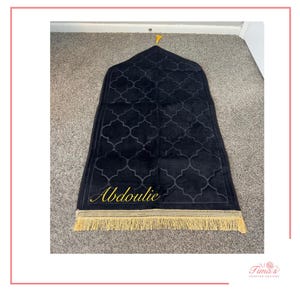 May include: A black prayer rug featuring a repeating quatrefoil pattern, a pointed arch top, and a gold-colored fringe at the bottom. The name Abdoulie is embroidered in gold script on the lower left side of the rug, with a small gold tassel.