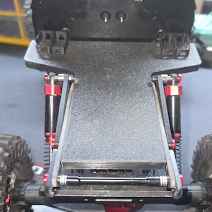 May include: Black RC car chassis with red and black suspension components. The chassis has a textured surface and features the "REDS" logo and "WARN" sticker. The car has black wheels and tires, and the "Axial" logo is visible.