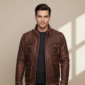 May include: Brown leather jacket with four front pockets and a collared neckline. The jacket has a distressed appearance and a zipper closure. The model is wearing a dark gray shirt underneath.