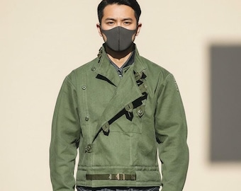 May include: A green jacket with a unique asymmetrical design, featuring a wrap-over front secured with straps and buttons. The jacket has a belt at the waist and is worn with a gray face mask and blue jeans. The jacket is a military-inspired style.