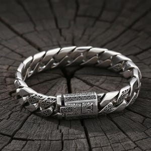May include: A silver bracelet with a curb chain design. The bracelet has an ornate clasp with engraved detailing. The links are thick and polished, reflecting light. The bracelet is displayed on a dark, textured wooden surface.