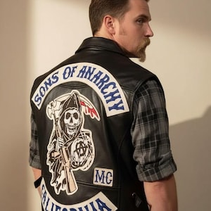 Puede incluir: Chaleco de cuero negro con el parche del logo "Sons of Anarchy" en la espalda. El parche presenta una parca con una guadaña y la palabra "California". El chaleco se lleva sobre una camisa de cuadros gris.