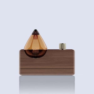 May include: A modern table lamp featuring a teardrop-shaped amber glass shade with a visible filament bulb. The lamp sits on a rectangular wooden base with a gold-colored knob. The design is minimalist and elegant.
