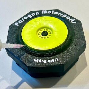 May include: A black octagonal tire holder with the text "Paragon Motorsports" and "1/8th Buggy" printed in white. A yellow tire is inside the holder. A syringe is injecting glue into the tire.