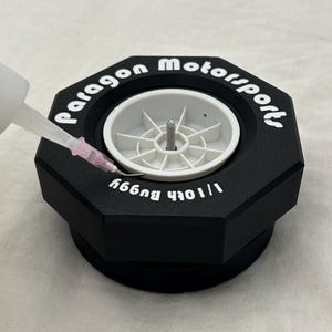 May include: A black octagonal tire stand with the words "Paragon Motorsports" printed in white. The stand has a white center with a small metal screw. A clear plastic syringe is dispensing liquid into the stand.