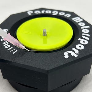 May include: A black, hexagonal, 3D printed base with the text "Paragon Motorsports" printed on the side. A yellow disc with a small metal screw in the center sits on top of the base.