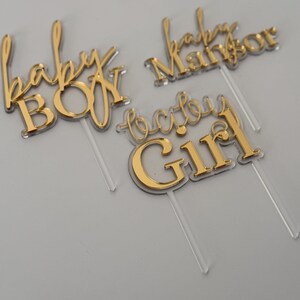 May include: Gold acrylic cake toppers with the phrases "baby BOY", "baby Manor", and "baby Girl". The text is in a cursive font with a clear plastic stick attached for insertion into a cake. The toppers are for a baby shower or gender reveal party.