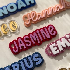 May include: A collection of custom name tags featuring various colours and textures. The names NOAH, Hannah, Leo, JASMINE, EMILY, MARIUS, and R are displayed in different fonts, including block letters and script, with layered acrylic and glitter finishes.