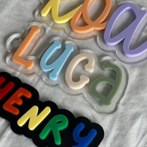 May include: Colorful acrylic name cutouts in various pastel colors, including yellow, orange, blue, and green. The names are mounted on clear cloud-shaped bases. One cutout spells "Henry" in rainbow colors on a black base.