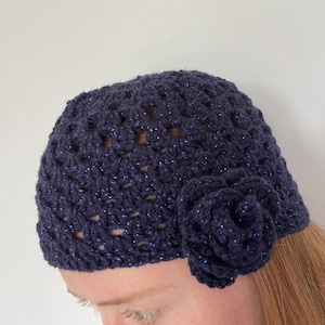 May include: A navy blue crochet hat with a sparkly thread woven throughout. The hat features a large, matching crochet flower on the side. The hat is a handmade accessory.