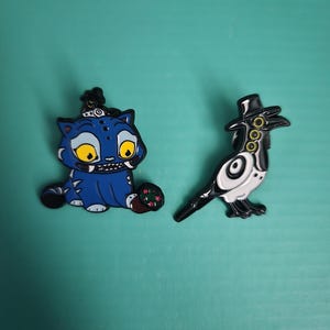May include: Two enamel pins: a blue cat with yellow eyes and a black and white bird wearing a top hat. The cat pin has a black crown and a red and green design on its paws. The bird pin has a black beak and feet.
