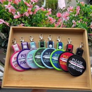 May include: A collection of eight circular embroidered keychains with silver-tone metal lobster clasps. Each keychain features a monorail train graphic and the text "PLEASE STAND CLEAR" in white embroidery, available in pink, purple, light blue, green, navy, lime, red, and black.
