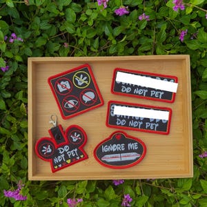 May include: A collection of four embroidered patches and a keychain with red borders and black backgrounds. The items feature white text including DO NOT PET and IGNORE ME, along with icons of a camera, a hand, an eye, and a speech bubble, all crossed out.