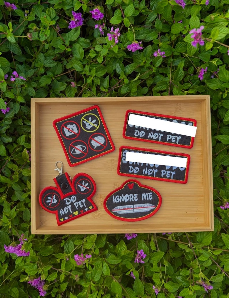 May include: A collection of four embroidered patches and a keychain with red borders and black backgrounds. The items feature white text including DO NOT PET and IGNORE ME, along with icons of a camera, a hand, an eye, and a speech bubble, all crossed out.