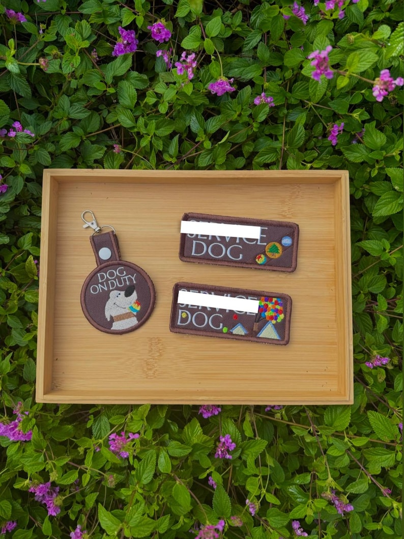 May include: A wooden tray holds three brown patches and a keychain. The patches read "SERVICE DOG" and "DOG ON DUTY", with colorful graphics. The keychain has a round patch with a dog graphic. The background is green foliage with purple flowers.