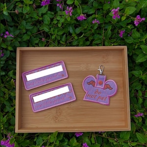 May include: Two rectangular embroidered patches with pink borders and the text one little spark, alongside a purple embroidered keychain shaped like a character head with horns and the text do not pet, featuring a silver metal lobster clasp.