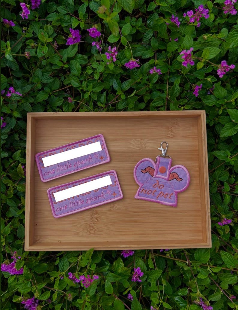 May include: Two rectangular embroidered patches with pink borders and the text one little spark, alongside a purple embroidered keychain shaped like a character head with horns and the text do not pet, featuring a silver metal lobster clasp.