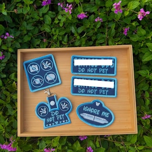 May include: A collection of dark blue embroidered patches and a keychain with light blue borders. The items feature icons of a camera, a hand, an eye, and a speech bubble with lines through them, along with the text DO NOT PET and IGNORE ME.