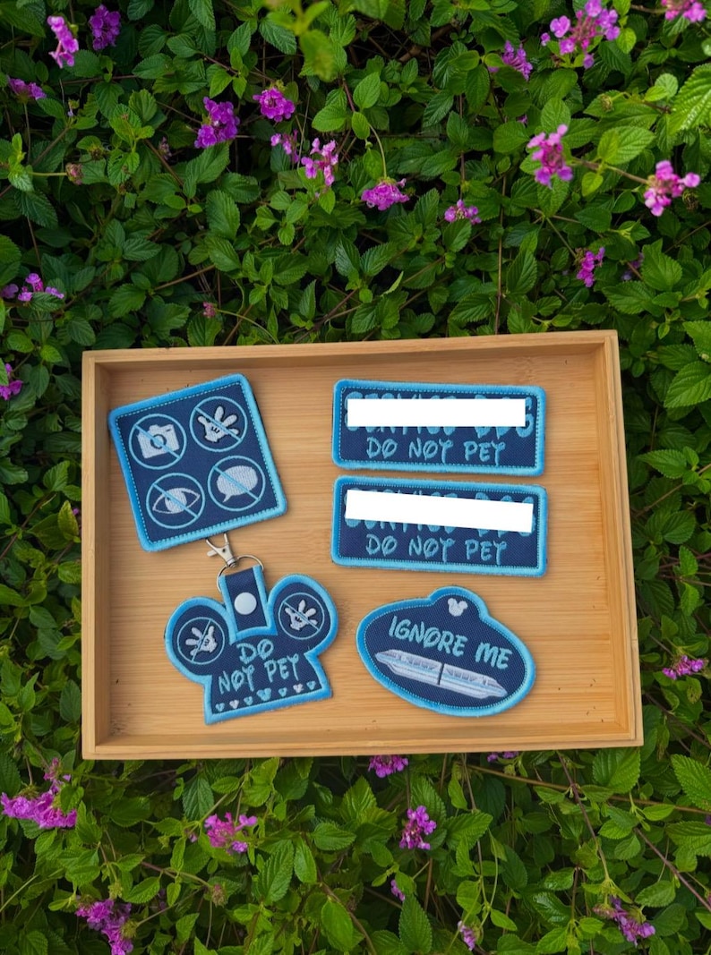 May include: A collection of dark blue embroidered patches and a keychain with light blue borders. The items feature icons of a camera, a hand, an eye, and a speech bubble with lines through them, along with the text DO NOT PET and IGNORE ME.