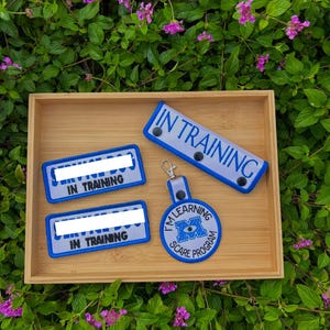 May include: Two rectangular patches with blue borders and white text reading SERVICE DOG IN TRAINING, one long rectangular strap with blue borders and text reading IN TRAINING, and a circular keychain with blue text reading I'M LEARNING SCARE PROGRAM.
