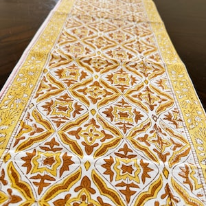 May include: A long, rectangular table runner featuring a repeating geometric pattern in shades of mustard yellow, burnt orange, and white. The fabric has a decorative border with a floral vine motif running along both of the long edges.