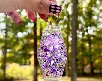 May include: A clear, motel-style keychain filled with purple glitter, stars, and a flower. The keychain has a gold ring and is held up by a hand with a black and pink checkered nail design. The background is a blurred view of trees.
