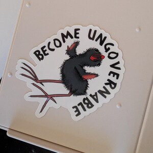 May include: A white sticker featuring the text "BECOME UNGOVERNABLE" around a cartoon bird-like character. The character is black with red details and long legs. The sticker is placed on a light beige surface.