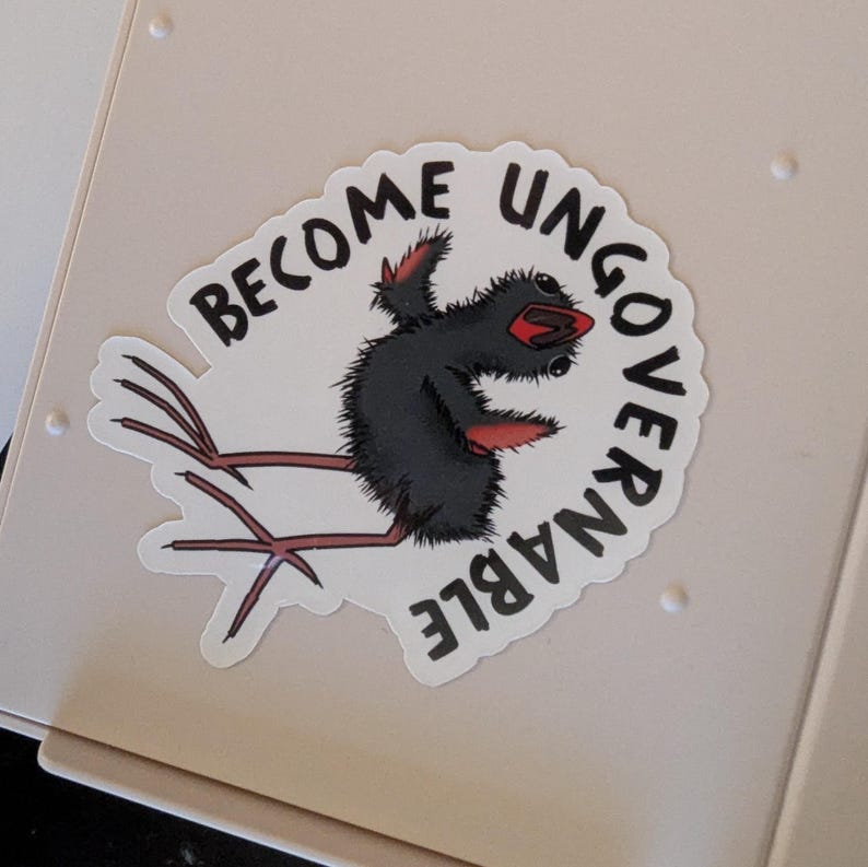 May include: A white sticker featuring the text "BECOME UNGOVERNABLE" around a cartoon bird-like character. The character is black with red details and long legs. The sticker is placed on a light beige surface.