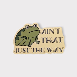 May include: A sticker featuring a cartoon frog with the text "AIN'T THAT JUST THE WAY" in black lettering. The frog is green with darker green markings, and the background is a light yellow.