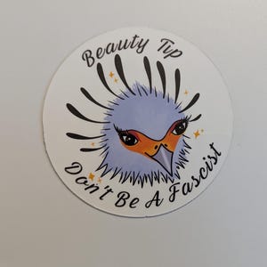 以下が含まれることがあります： スタイリッシュな鳥の顔が描かれた丸いステッカー。薄い青色の頭、オレンジ色のくちばし、長い黒いまつげが特徴です。「Beauty Tip」の文字が鳥の上に、「Don't Be A Fascist」の文字が下に弧を描いています。
