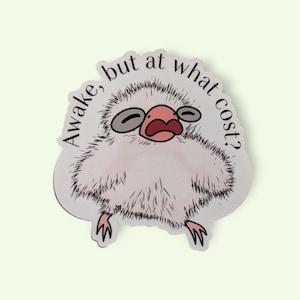 May include: A sticker featuring a cartoon illustration of a fluffy, light-colored creature with a pink face and open mouth. The text "Awake, but at what cost?" is printed around the creature's head in a semi-circular fashion.