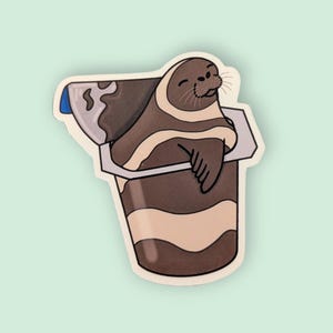 May include: A sticker featuring a cartoon seal relaxing in a chocolate pudding cup. The seal is brown and tan, with a smiling face. The pudding cup is brown and beige, with a blue lid. The sticker has a white border and a light green background.