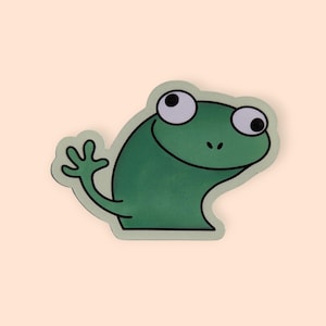 May include: A green frog sticker with a friendly expression, waving a hand. The frog has large white eyes with black pupils and a light green outline. The sticker is on a light peach background.