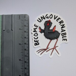 May include: A sticker featuring a black bird illustration with red accents and long legs. The sticker has the words "BECOME UNGOVERNABLE" in a semi-circle. The sticker is approximately 7.6 cm in height.