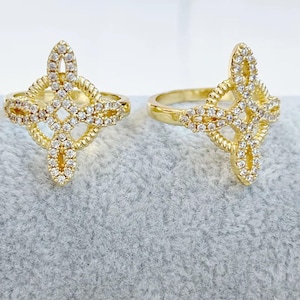 May include: Two gold-colored rings with a diamond-studded, cross-shaped design. The rings feature a textured band and a central diamond pattern. The rings are displayed on a gray surface.