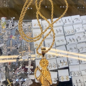 May include: A gold-toned rope chain necklace featuring a pendant shaped like a figure holding a scythe. The pendant depicts a robed entity with a halo, rendered in a polished metallic finish. The necklace is displayed with a lobster claw clasp.